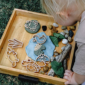 Child playing with nature-themed playdough and materials on a wooden tray outdoors.