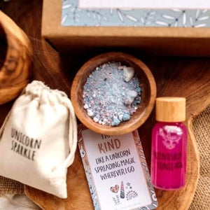 A styled flat-lay of a magic potion gift set on a wooden tray, featuring a small cream drawstring pouch labeled 'UNICORN SPARKLE', a wooden bowl of pastel salts, a pink bottle labeled 'RAINBOW TONIC', and an affirmation card.