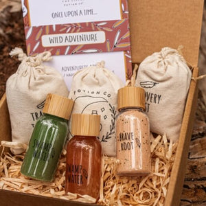 A boxed magic potion kit from The Little Potion Co featuring three small bottles with bamboo caps containing coloured ingredients and labeled "Swamp Water", and "Brave Root", three beige drawstring pouches, and a bundle of printed cards inside a cardboard box filled with shredded paper.