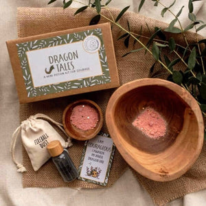 A styled flat-lay of the Dragon Tales Mini Potion Kit for Courage, featuring a kraft paper box with a green leaf border, a wooden mortar and pestle containing pink powder, a small wooden bowl with pink powder, a cream drawstring pouch labeled 'Fireball Dust', and a small bottle with a wooden cap and card.
